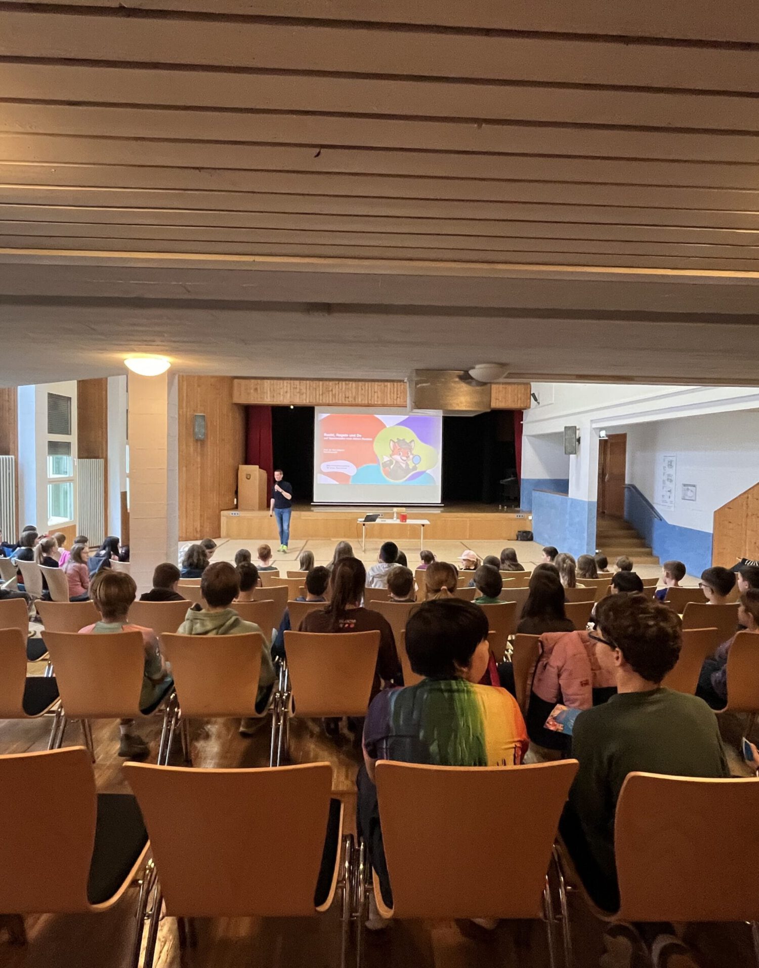 Aula der Kinderuniversität mit sitzenden Kindern im Publikum und einer laufenden Präsentation vorne.